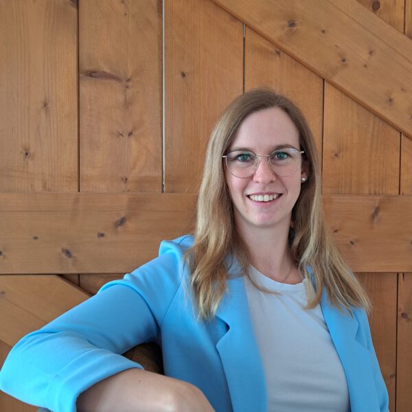 Traurednerin Vanessa Grasser sitzt auf einem Stuhl vor einem Holztor und lächelt in die Kamera. Worte mit Herz - freie Trauung Kontakt-Formular