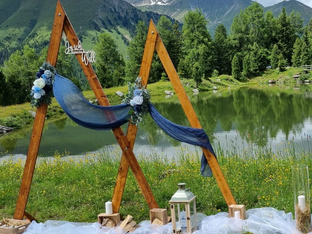 Traubogen aus Holz von einer Berghochzeit Destinationwedding in Tirol mit Bergsee im Hintergrund von Traurednerin Vanessa Grasser